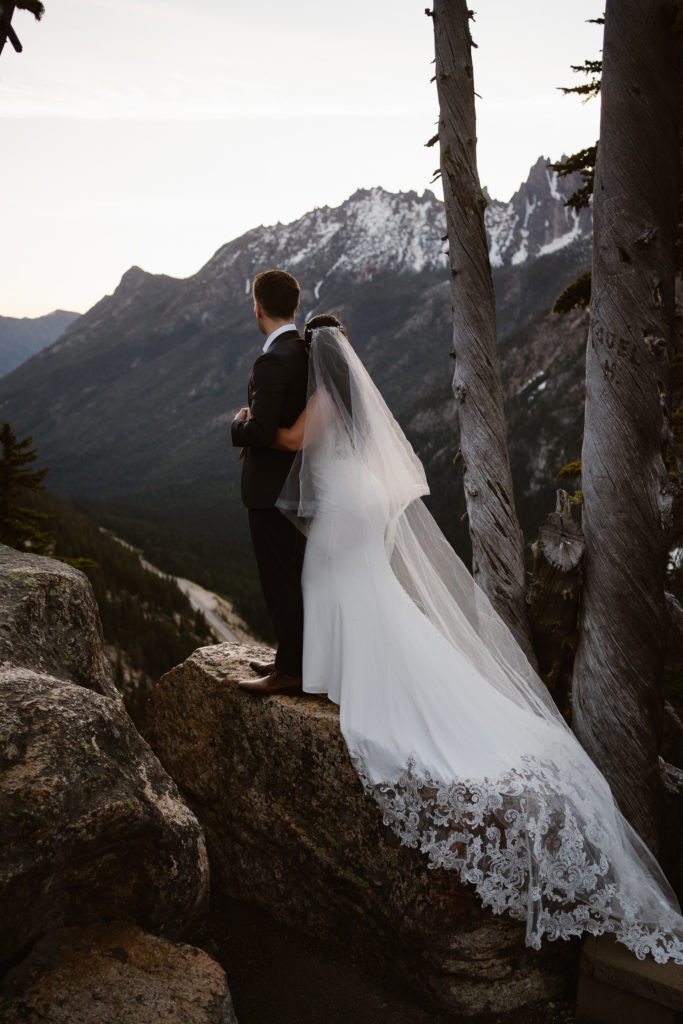 a bride hugs a groom from behind as they stand on a rock above a cliff