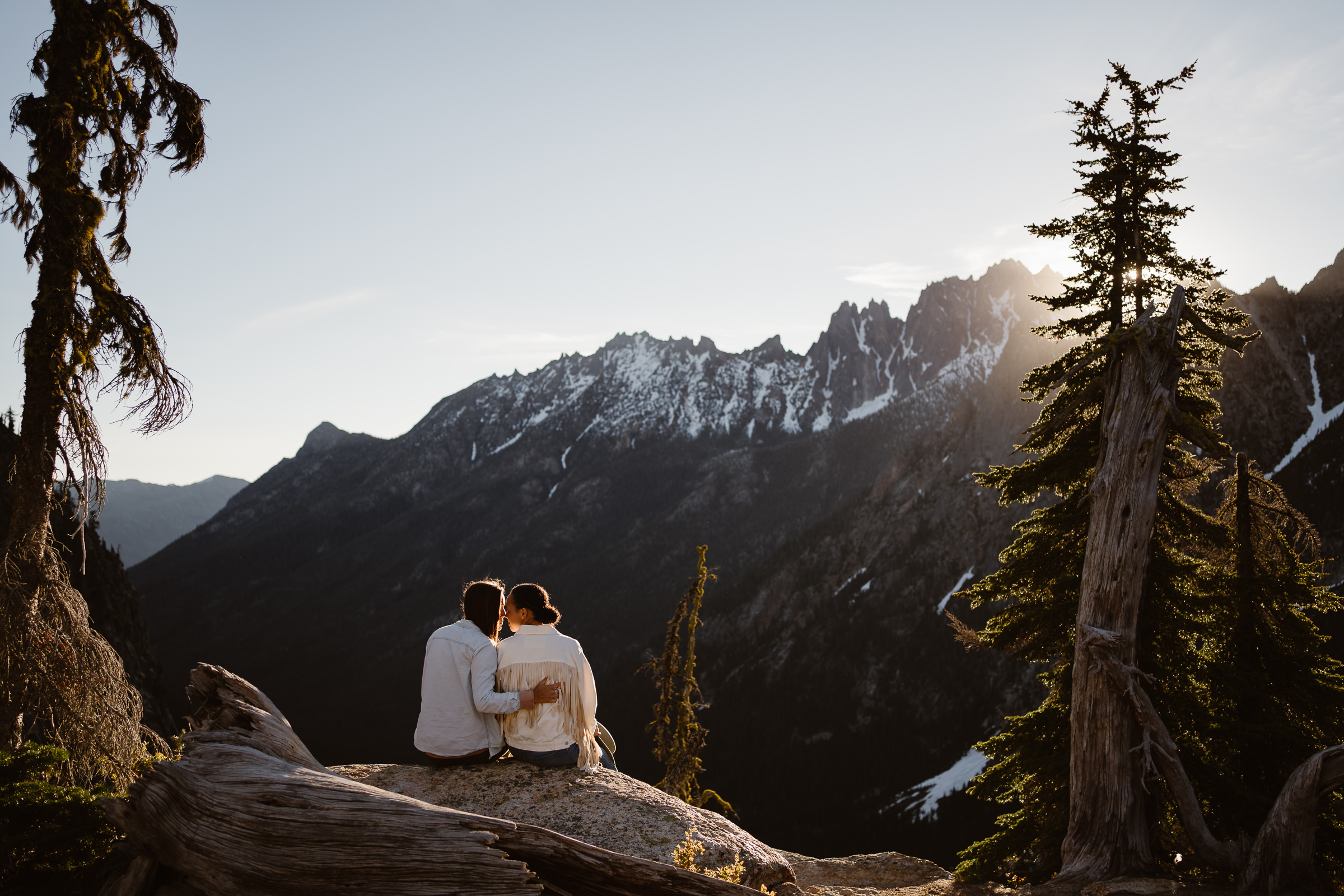 a couples sits on a log and watches the sun rise over the mountain ridge in front of them during their north cascades engagement session