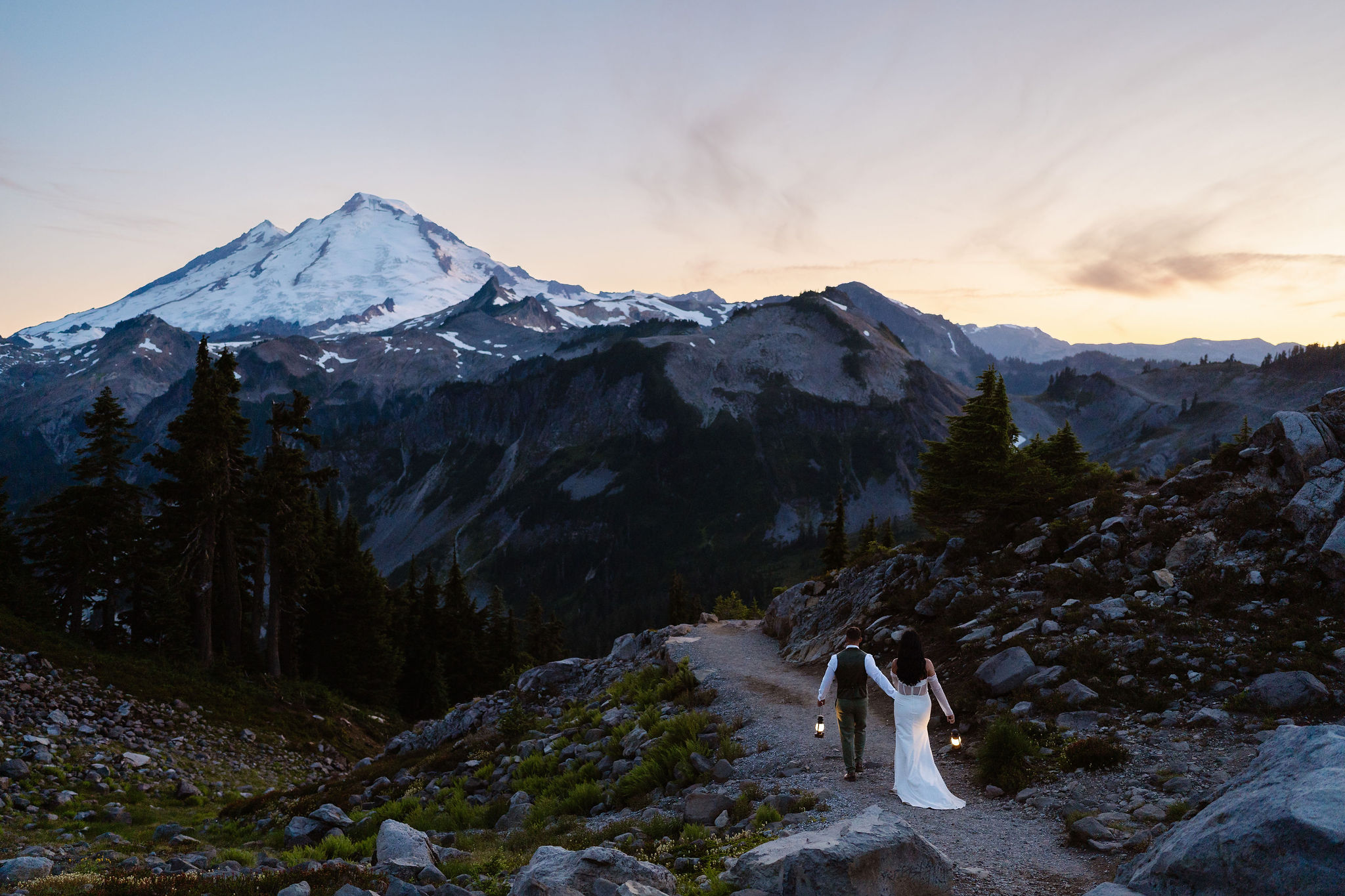 an eloping couple holds hands and lanterns as they walk off into the sunset at golden hour towards mt baker during their north cascades elopement