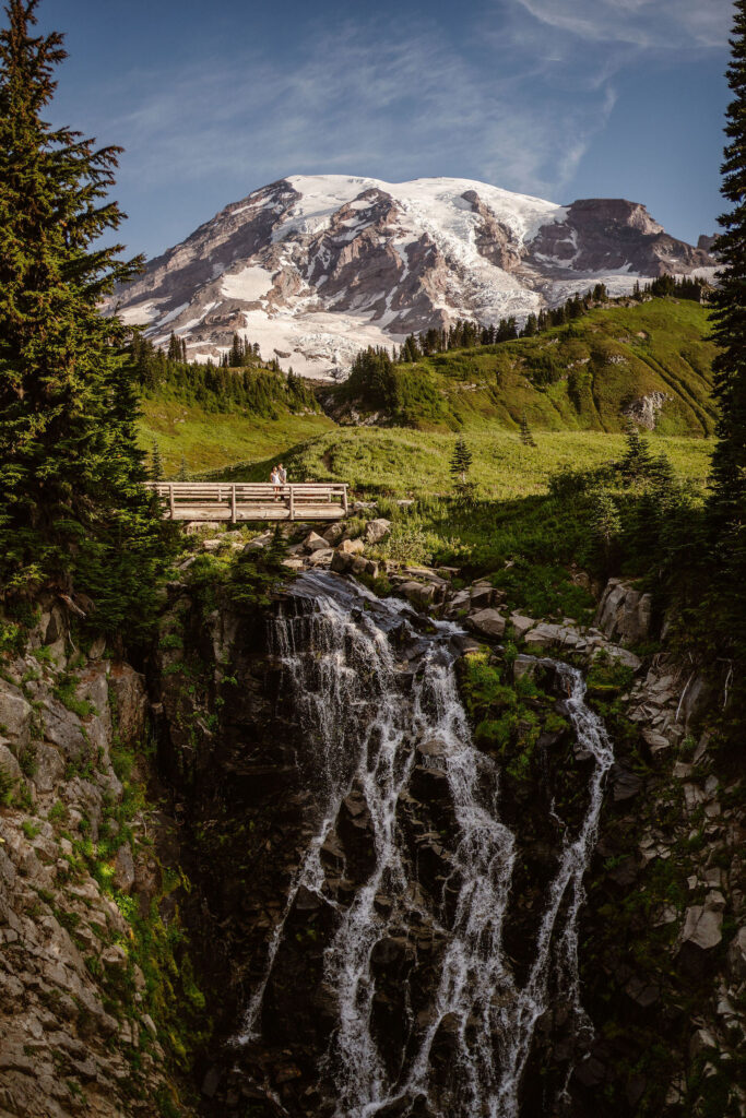 a couple stands on a bridge over a waterfall with Mount Rainier in the background