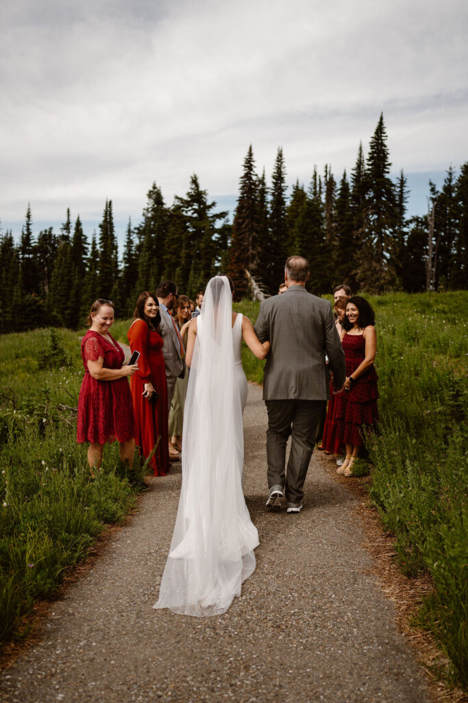 a bride is walked down the aisle by her dad as guests stand on either side of the trail during a mt rainier micro wedding