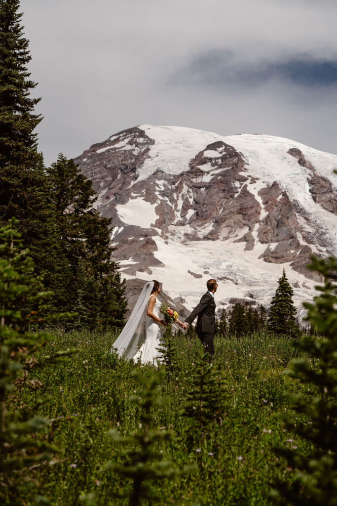 a bride and groom hold hands and walk down the trail with mount rainier in the background during their mt rainier micro wedding