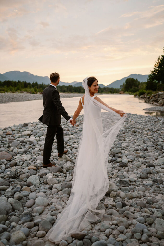 a bride and groom hold hands while the bride plays with her veil along the cowlitz river