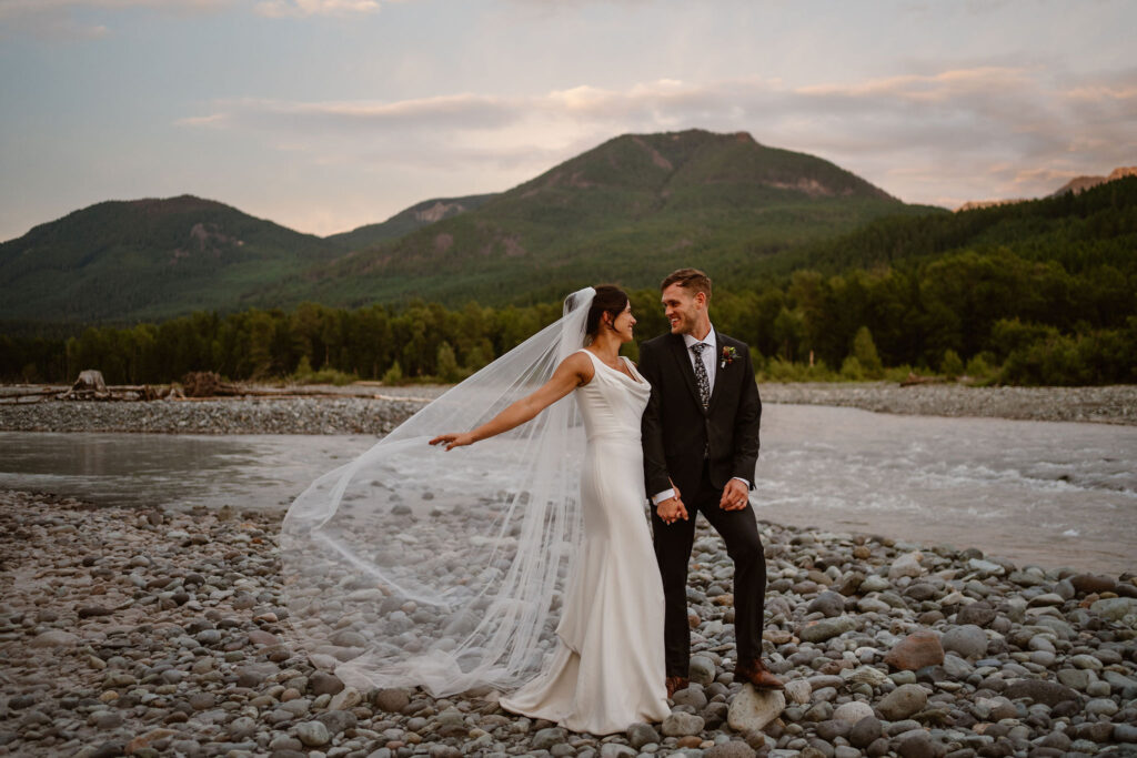 a bride and groom hold hands and smile at each other while the bride plays with her veil in the wind